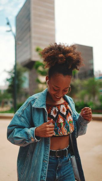 Closeup, smiling African girl with ponytail wearing denim jacket, in crop top with national pattern listening to music on headphones and dancing outdoors.  - Photo, image