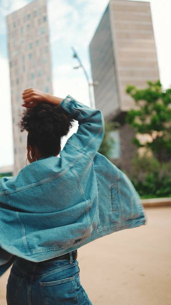 Closeup, smiling African girl with ponytail wearing denim jacket, in crop top with national pattern listening to music on headphones and dancing outdoors.  - Φωτογραφία, εικόνα