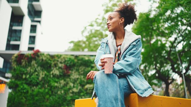 Cute African girl with ponytail wearing denim jacket, in crop top with national pattern, drinks morning coffee on modern city background. - 写真・画像
