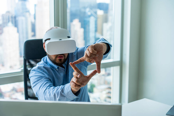 Serious bearded male entrepreneur in VR goggles showing frame while watching augmented world in modern workplace in office with panoramic windows - Φωτογραφία, εικόνα