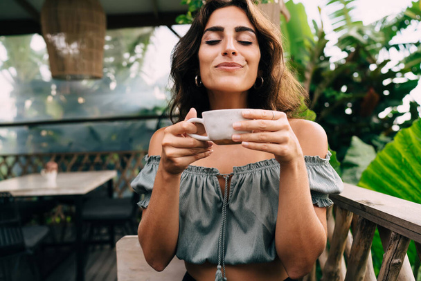 Delighted young ethnic lady with long hair sitting with eyes closed and enjoying flavor of hot coffee on balcony of modern cafeteria - Foto, imagen