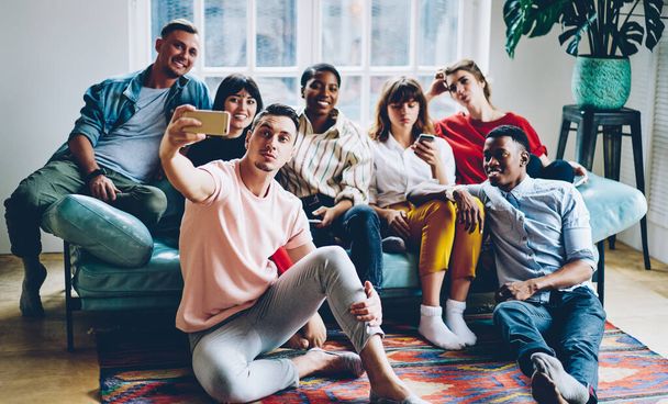 High angle of joyful millennial multiethnic students in vivid casual clothes taking selfie with smartphone during meeting in contemporary apartment in free time - Photo, Image