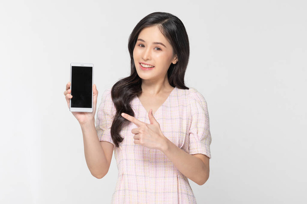 Happy attractive asian young woman smile and showing mobile phone isolated on white background. Cheerful pretty girl wear casual dress raise up smartphone to empty space for mockup to display product - Valokuva, kuva