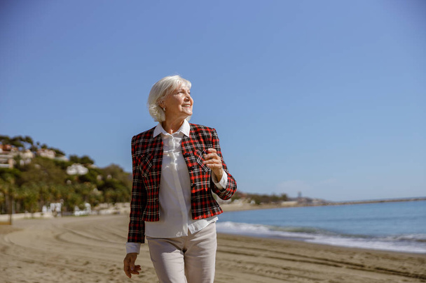 Good looking senior woman wearing plaid jacket walking outdoors against backdrop of picturesque seascape - Photo, Image