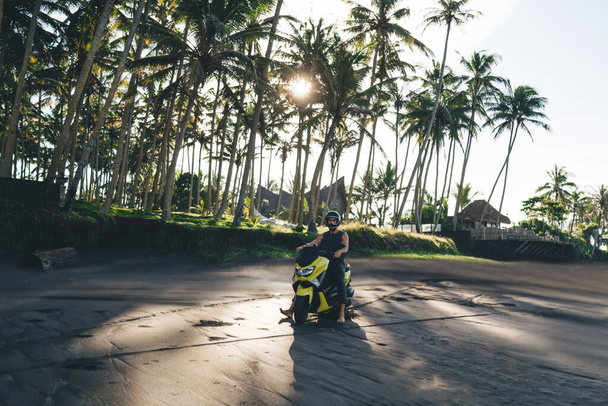 Full length of unrecognizable male biker wearing protective helmet driving motorcycle along empty sandy beach near palm trees on sunny day - Foto, imagen