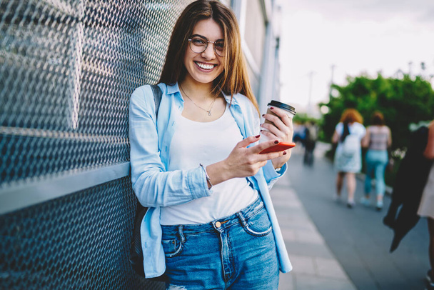 Half length portrait of cheerful hipster girl with smartphone for online blogging smiling at camera and rejoicing in city - Foto, Imagen