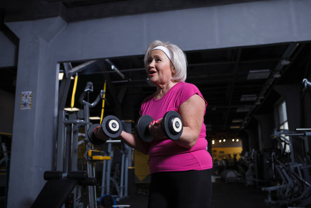 Veselý senior žena těší cvičení v tělocvičně, dělat biceps kadeře s činkami - Fotografie, Obrázek