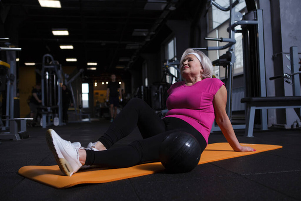 Relaxed elderly woman sitting on yoga mat, resting after workout at gym - Fotó, kép