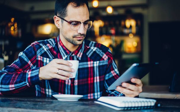 アメリカーノコーヒーを飲みながらスタイリッシュなメガネを持つビジネスマンが、カフェテリアのインテリアで無料の時間の間にデジタルタブレットでウェブニュースを読む - 写真・画像