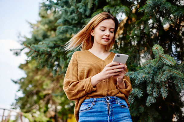 Von unten eine fokussierte junge Frau in Jeans und Strickjacke, die mit einem Freund auf dem Smartphone SMS schreibt, während sie in der Nähe von Nadelbäumen im Park steht - Foto, Bild