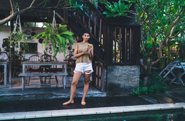 Full body optimistic ethnic female in casual clothes standing on poolside of hotel area surrounded by plants in Bali while using smartphone alone - Foto, Bild