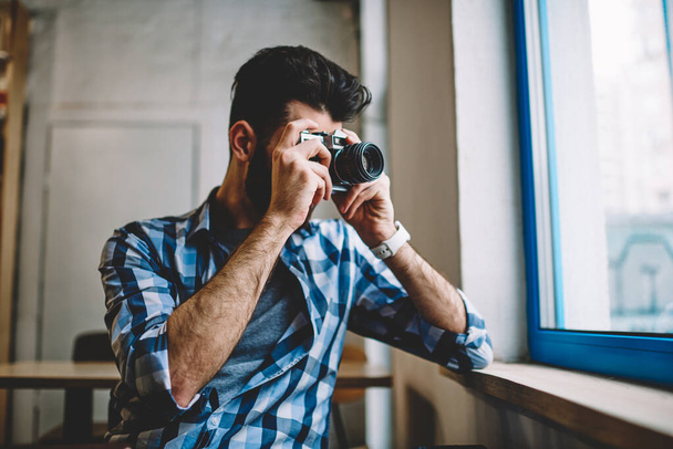 Talented creative caucasian hipster guy making pictures using vintage camera near cafeteria window, young professional male photographer taking images via retro old fashioned technology indoors - 写真・画像