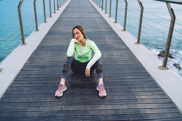 High angle of positive middle age female in sportswear and sneakers resting on wooden pier after workout and looking at camera - Фото, изображение
