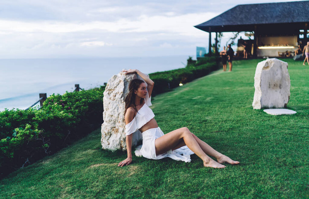High angle of full body relaxed barefoot slim female in elegant white dress resting on green grass and looking away - 写真・画像