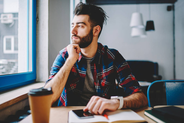 Thoughtful young man dressed in casual wear looking away while sitting at desktop with coffee to go in university.Pondering bearded hipster guy thinking on ideas for development of own projec - Foto, imagen