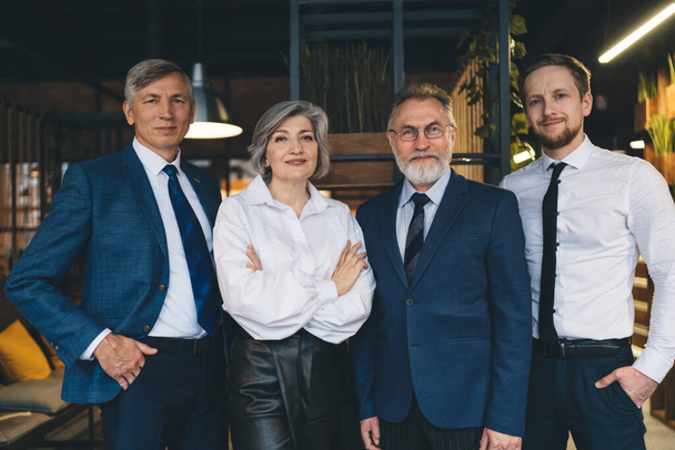 Confident mature woman and men in suits standing together in modern office workplace with modern interior and looking at camera - Фото, зображення