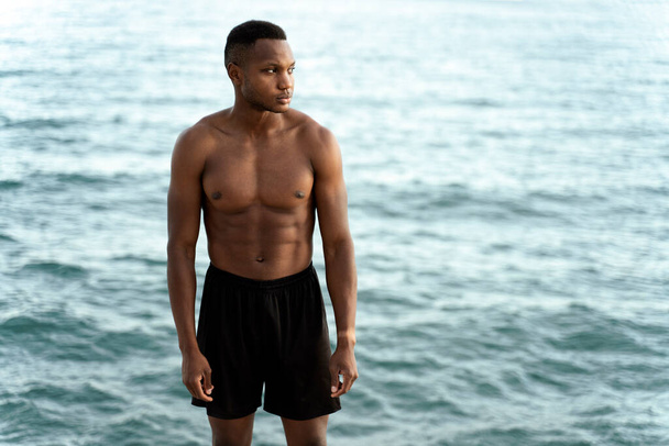 Αφροαμερικάνος με σπορ σώμα και σε καλή κατάσταση κοιτάζει αλλού και συλλογίζεται ενώ στέκεται στην παραλία με κυματιστή θάλασσα στο βάθος - Φωτογραφία, εικόνα