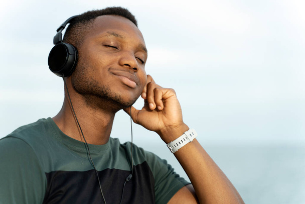 Close up portrait of calm handsome fitness athletic man in sport t-shirt listening music put hand on his ear while resting with closed eyes near the sea outdoors  - Φωτογραφία, εικόνα