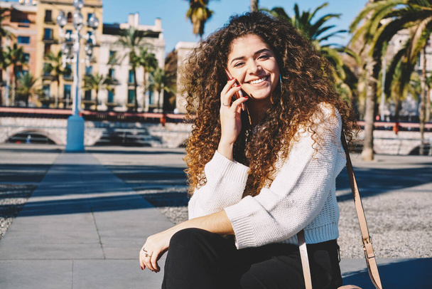 Portrait of cheerful female tourist with curly hair smiling at camera while using cellphone technology for friendly calling - Photo, image