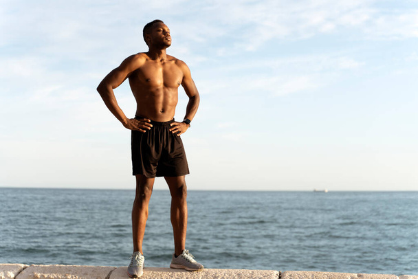 Ganzkörperansicht des afrikanisch-amerikanischen Mannes, der mit nacktem Oberkörper und sommerlichem Meer im Hintergrund posiert. Archivbild  - Foto, Bild