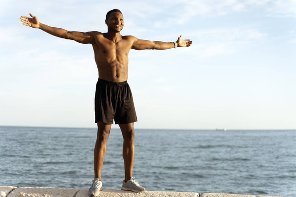 Full length view of african american man with naked torso celebrating victory or good results while standing near the sea  - Photo, Image