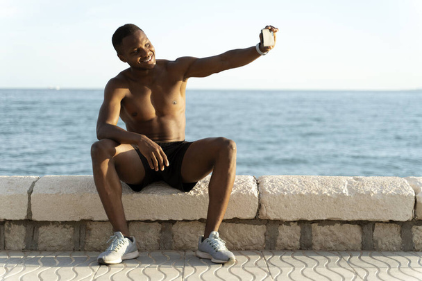 Jonge sterke multiraciale sportieve atleet fit sportman doen selfie schot op mobiele telefoon aan het strand buiten op pier kust in de zomer 's morgens  - Foto, afbeelding