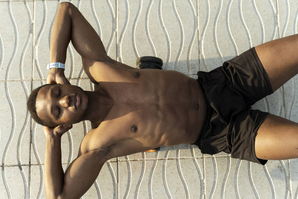 Bovenaanzicht van de sportman die buikspieroefeningen doet, fitnesstraining op straat. Multiraciale man met naakte romp die aan de kust traint  - Foto, afbeelding