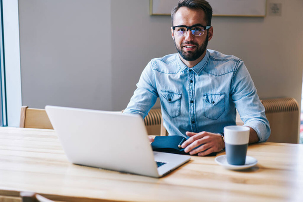 Bearded male freelancer in denim shirt looking at camera sitting at table with notebook and cup of tea while working on laptop - Foto, Imagen