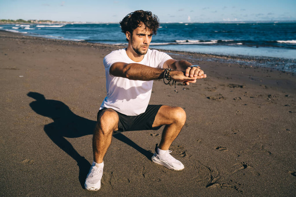 Pełne ciało brodatego mężczyzny biegającego w odzieży sportowej i tenisówkach robiącego przysiady przed joggingiem na wybrzeżu w letni dzień i odwracając wzrok - Zdjęcie, obraz