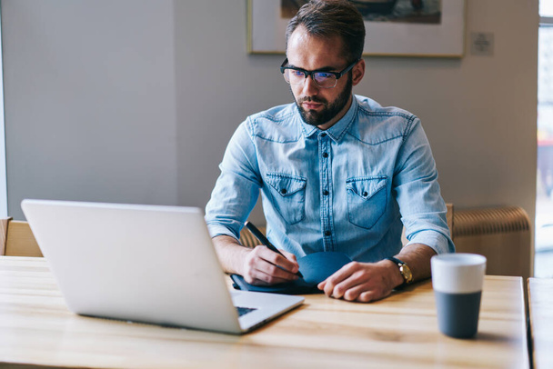Concentrated male freelancer in denim shirt looking at laptop taking notes while sitting at table with notebook and cup of coffee - Photo, Image