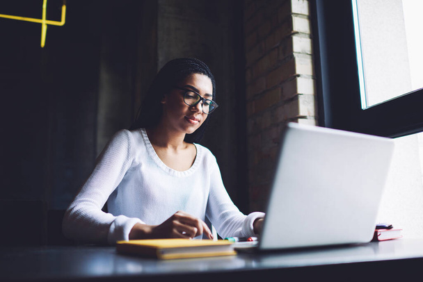 Tranquil African American female freelancer in eyeglasses using laptop while sitting at table with notebook near window in dark workspace - 写真・画像