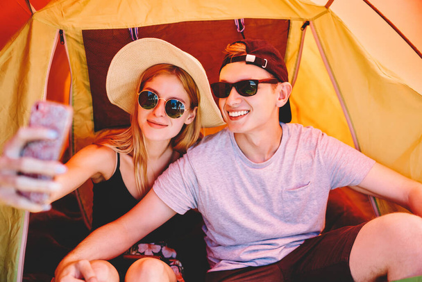 Cheerful tourists in summer casual outfit and sunglasses taking selfie together using mobile phone in camping tent on summer holidays - Fotó, kép