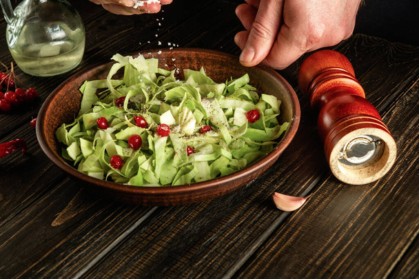 Profesionální kuchař sype sůl do salátu čerstvé zeleniny na dřevěný stůl. Příprava zdravého jídla v kuchyni - Fotografie, Obrázek