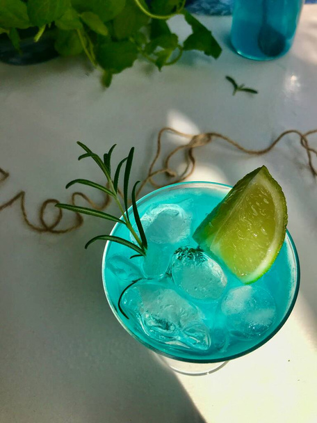 Голубой холодный напиток в стакане с кубиками льда на белом столе с голубым фоном. Также используются мятные листья и розмарин, веревка, ложка с натуальным солнечным светом из открытого окна - Фото, изображение