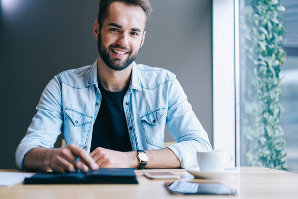 Positive entrepreneur in casual clothes sitting at table with notebook and cup of drink and looking at camera in light modern workspace - Φωτογραφία, εικόνα