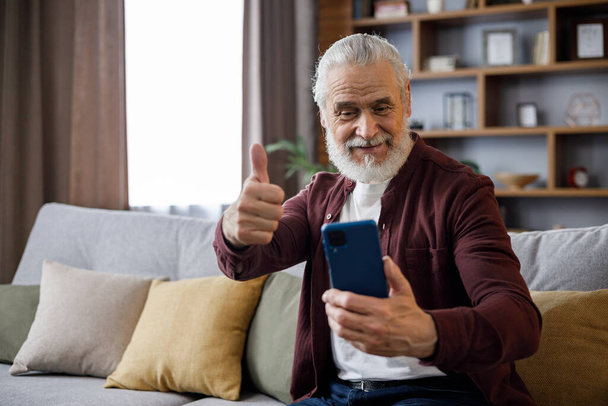 Smiling older man sitting on couch giving thumb up while playing computer game, looking at camera, smiling - 写真・画像
