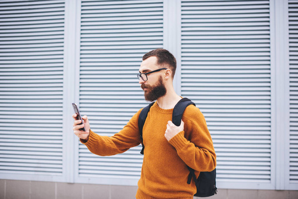 Side view of young male in trendy outfit and eyeglasses with backpack taking selfie on mobile phone beside building with louvers on windows - Fotoğraf, Görsel