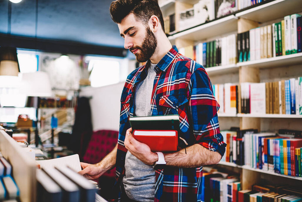 Pensive bearded hipster student choosing textbook from shelf in college for studying and writing coursework.Smart young man dressed in casual wear spending time in library of university - Foto, Imagen