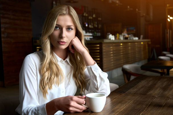 Mulher loira jovem atraente bebendo café no café moderno e pensando sobre a ideia de inicialização. Interior de madeira marrom. Foto de alta qualidade - Foto, Imagem