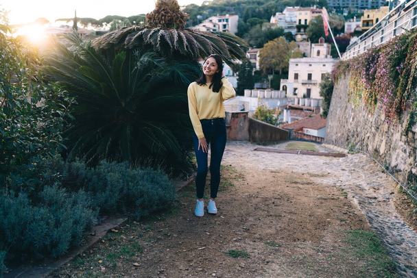Full length of cheerful ethnic female in casual clothes standing on ground with tropical bushes and smiling while looking at camera and posing - Φωτογραφία, εικόνα
