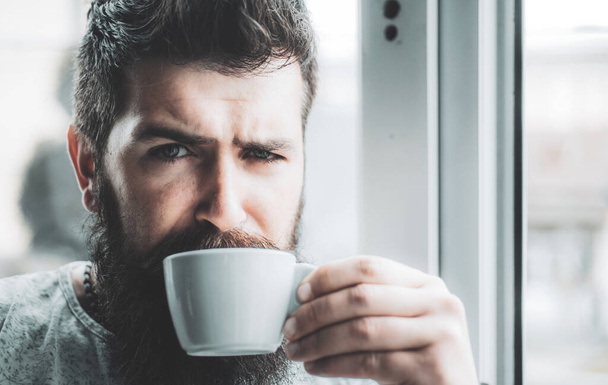 Grumpy looking bearded man drinking morning cup of coffee near window. Hipster man having cup of americano. Morning routine with coffee and start of new day. Coffee addicted - 写真・画像