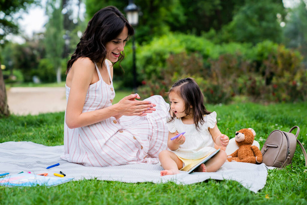 Side view of happy young woman sitting on blanket on green lawn with daughter drawing with color pencil and clapping hands while looking - Foto, immagini