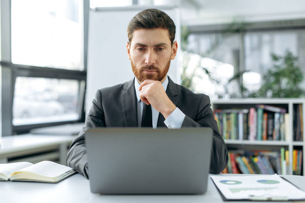 Сосредоточенный умный кавказский мужчина, маркетолог, веб-разработчик, финансовый менеджер, сидит за рабочим столом перед ноутбуком, в современном офисе в стильном деловом костюме, сконцентрированный на работе в ноутбуке - Фото, изображение