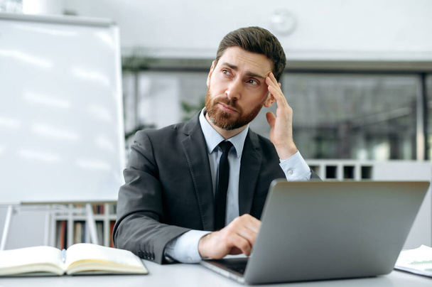 Страстный белый мужчина с бородой, директор компании, топ-менеджер, сидит за столом перед ноутбуком в современном офисе, задумчиво смотрит в сторону, анализирует рабочее задание, обдумывает стратегию - Фото, изображение
