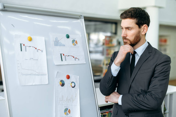 Focused elegant young adult caucasian man, company top manager, financial analyst, risk manager, stands near whiteboard with graphs in a modern office, concentrated looks at them, analyzes dynamics - Photo, Image