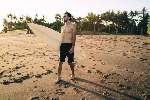 Full body of concentrated young bearded male athlete with naked torso standing on sandy beach with surfboard and admiring sunset in tropical resort - Fotó, kép