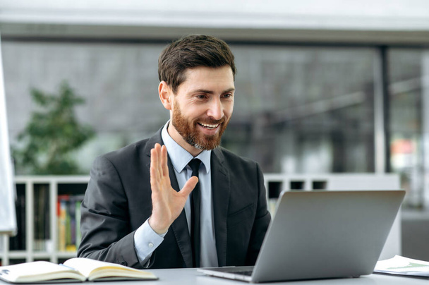 Prospective successful Caucasian male, lawyer, manager, economist, sits at a desk in modern office, wearing stylish business suit, holding video consultation with a client, waving a greeting, smile - Photo, image