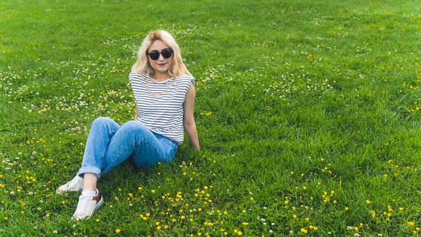 屋外でフルショット芝生の上でリラックスサングラスを持つブロンドのヨーロッパの女の子。高品質の写真 - 写真・画像