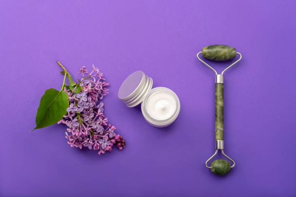 Jade facial roller, cream jar and lilac flower on purple background. Natural cosmetics, skin care concept. Top view, flat lay. - Fotoğraf, Görsel