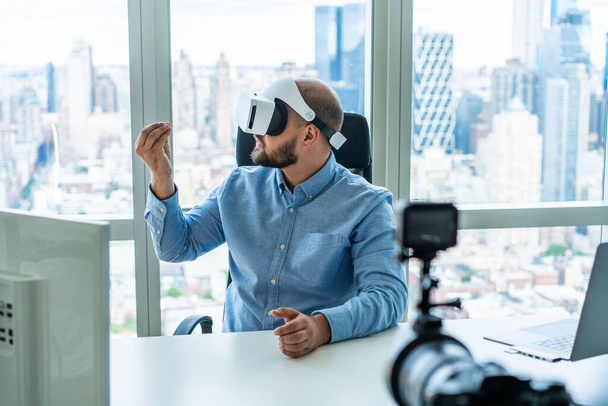 Concentrated male executive worker wearing VR goggles sitting at table with monitor laptop and recording video camera in contemporary workplace - Foto, Imagem